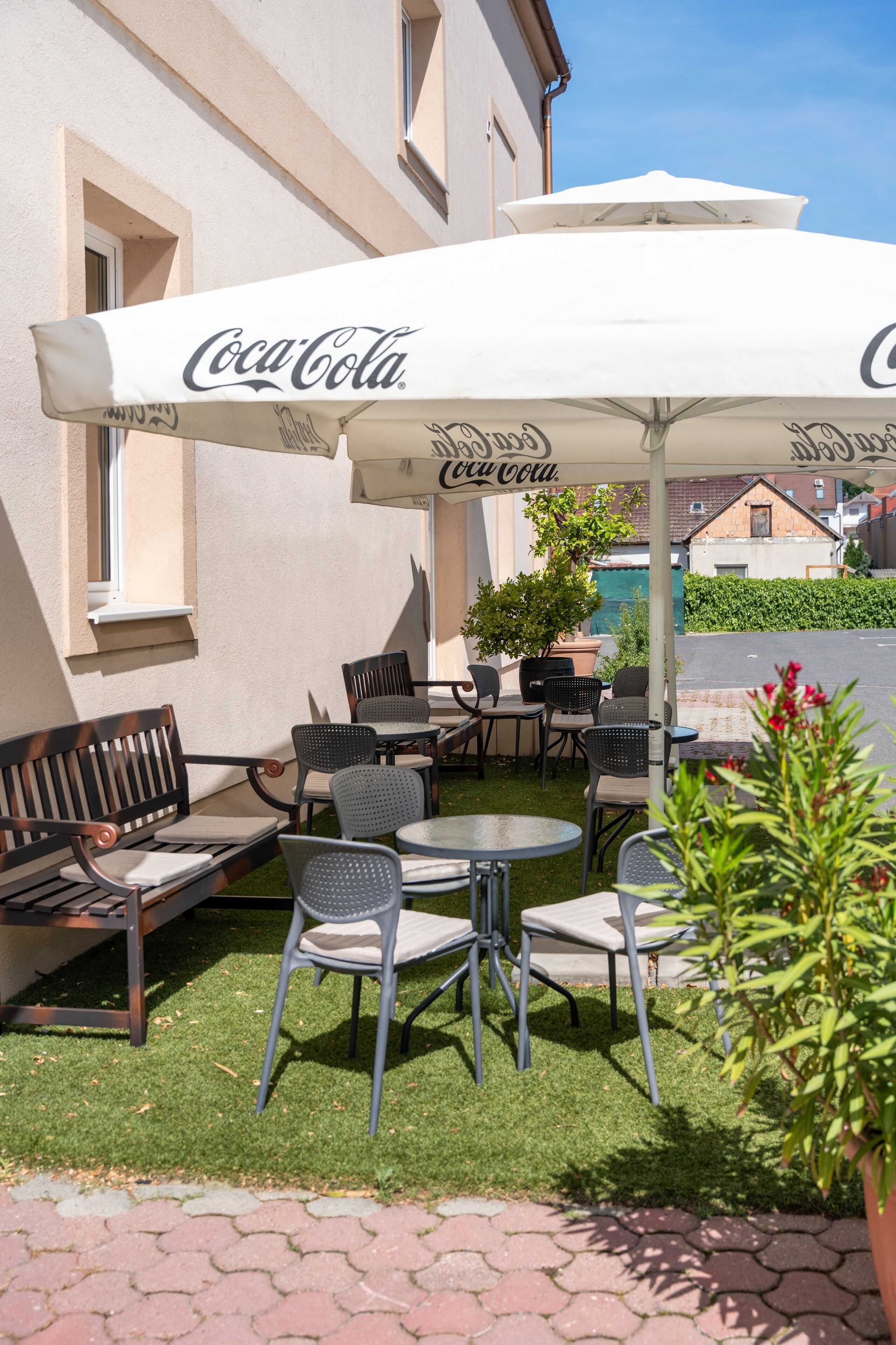 Un espace extérieur aménagé avec des tables et des chaises, protégé par un parasol à l’effigie de Coca-Cola, entouré de verdure et bordé d’un chemin pavé.