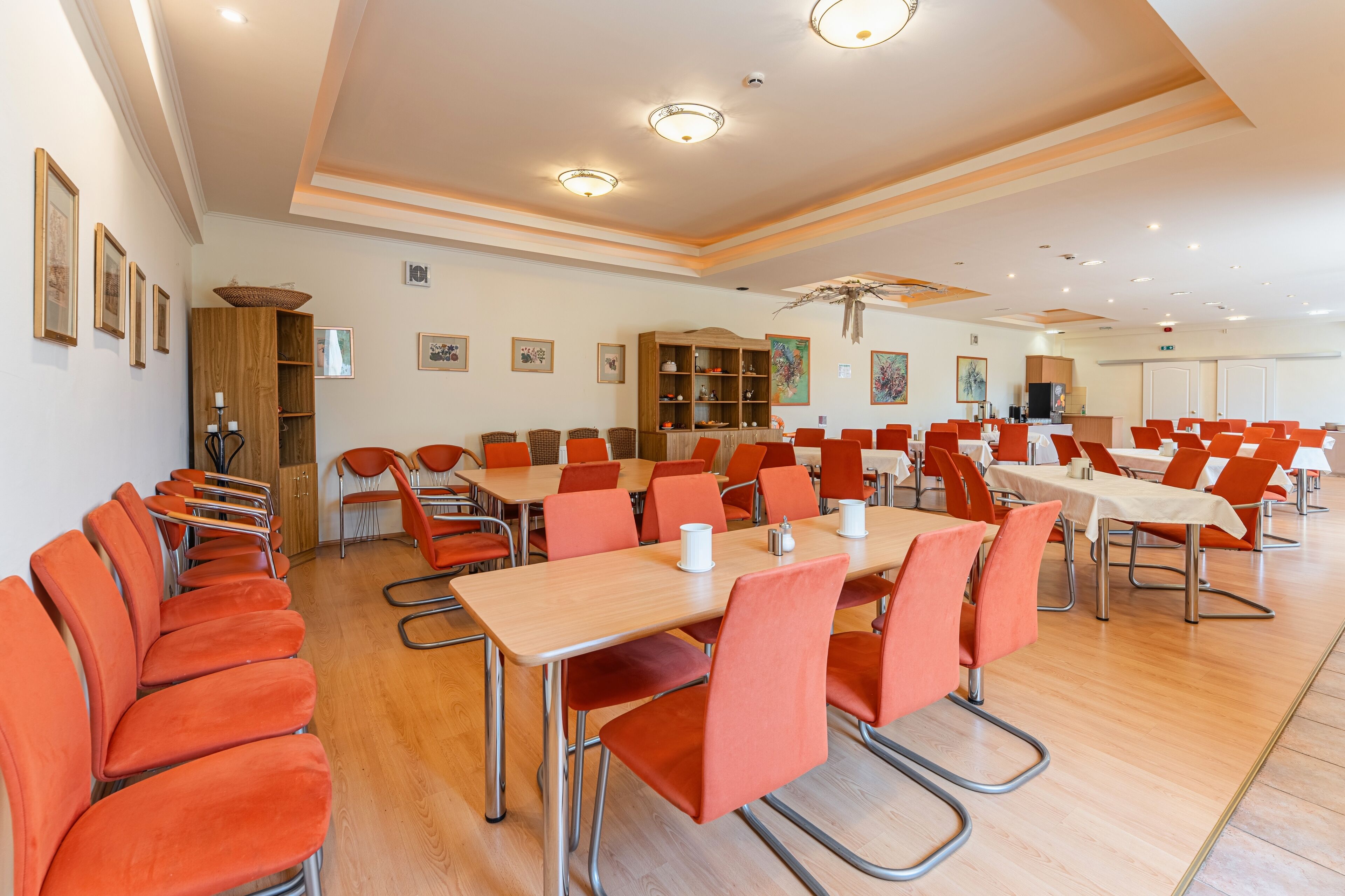 A spacious conference room with multiple tables and orange chairs, wooden cabinets, and framed artwork on the walls.
