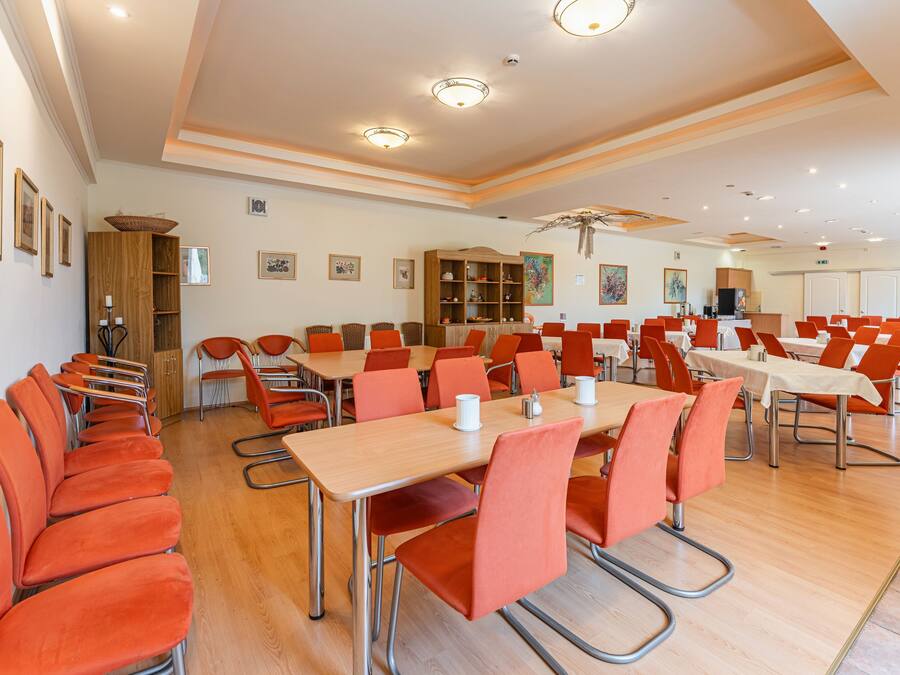 A spacious conference room with multiple tables and orange chairs, wooden cabinets, and framed artwork on the walls.