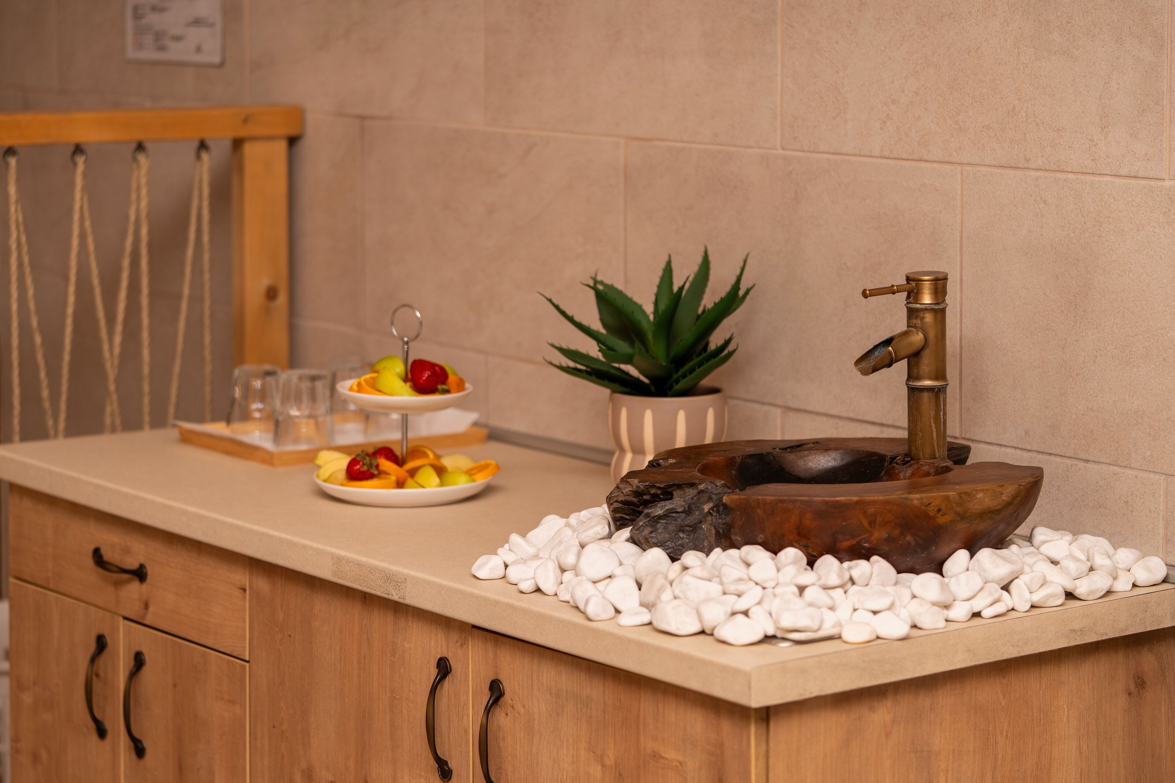 A kitchen counter with a wooden sink, white pebbles, a fruit platter, a potted plant, and a hanging rack.