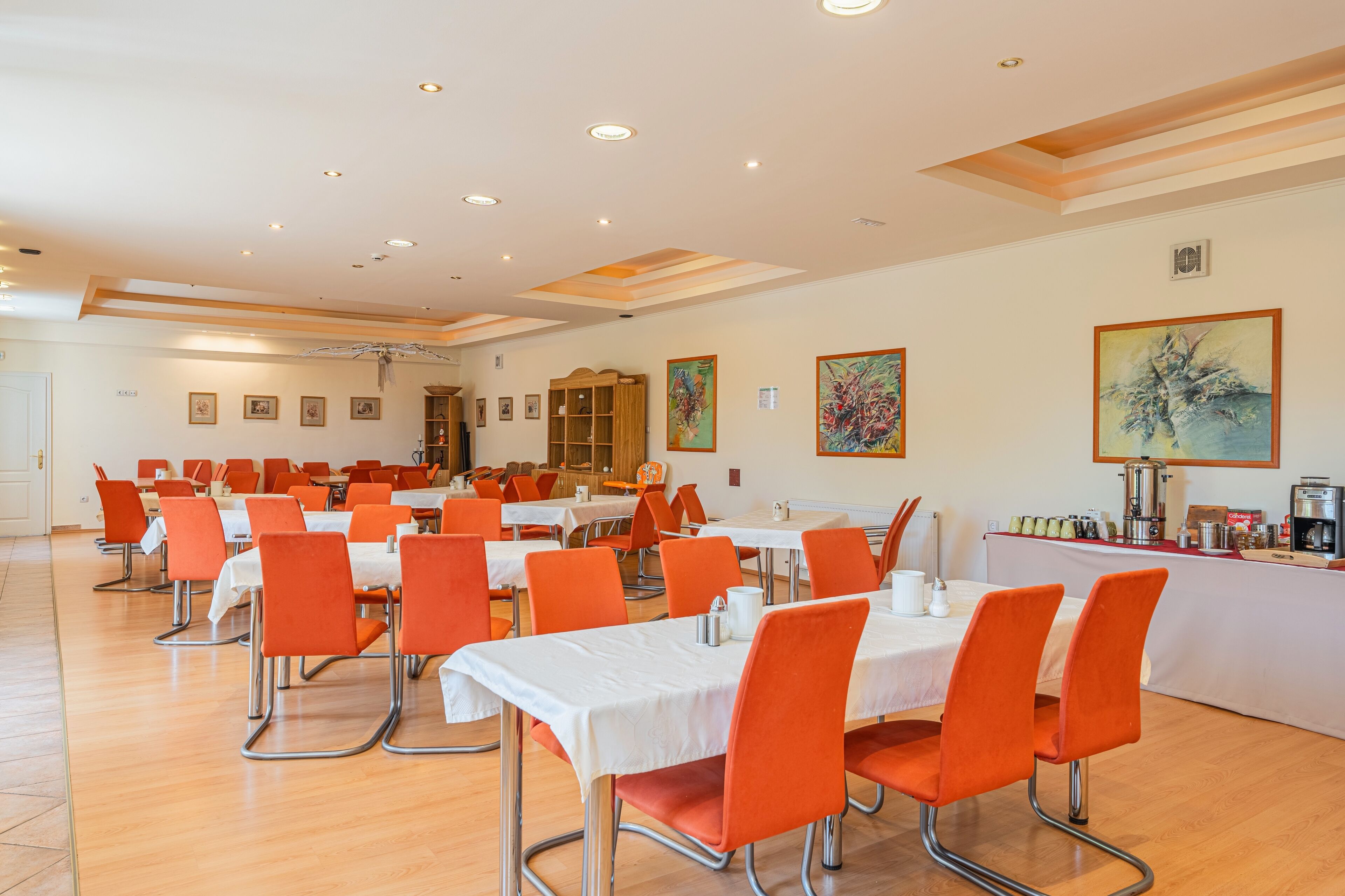 A spacious dining area with multiple tables and orange chairs, featuring a clean, modern design with white tablecloths and a wooden floor.