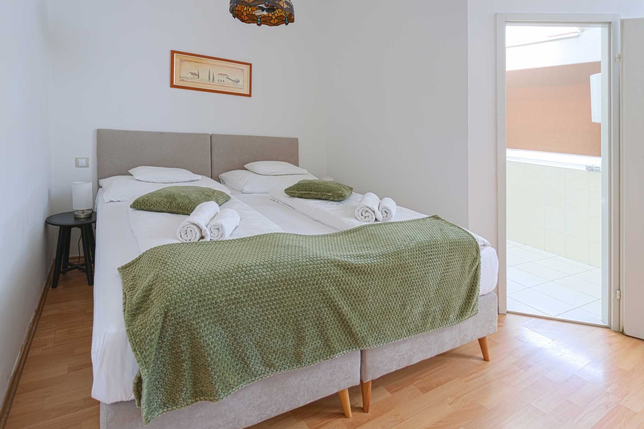 A bed with a green blanket, white pillows, and rolled towels.