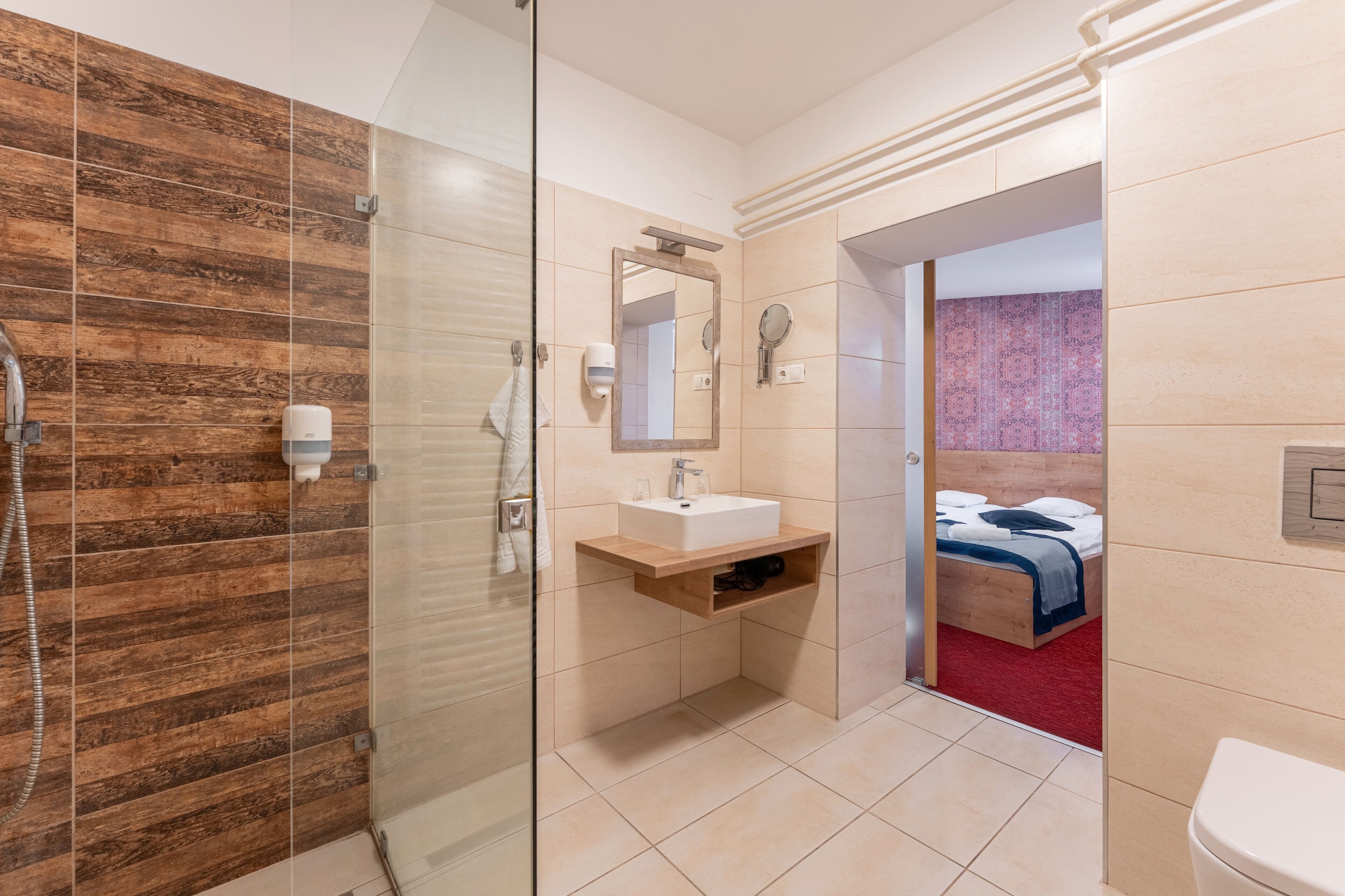 Ein modernes Badezimmer mit Duschkabine aus Glas, einem Holzbadezimmer-Möbel und einem Bett, das durch einen offenen Durchgang sichtbar ist.