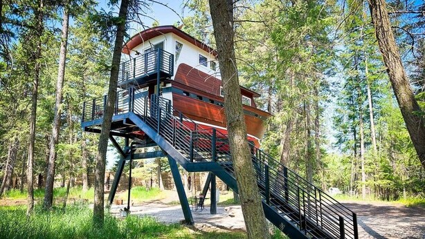 Une cabane en bois à plusieurs niveaux, dotée d’un escalier et d’un balcon, entourée d’arbres.