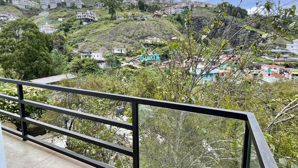A balcony with a view of a hillside village with houses and greenery. - The Lake House (Kodaikanal)