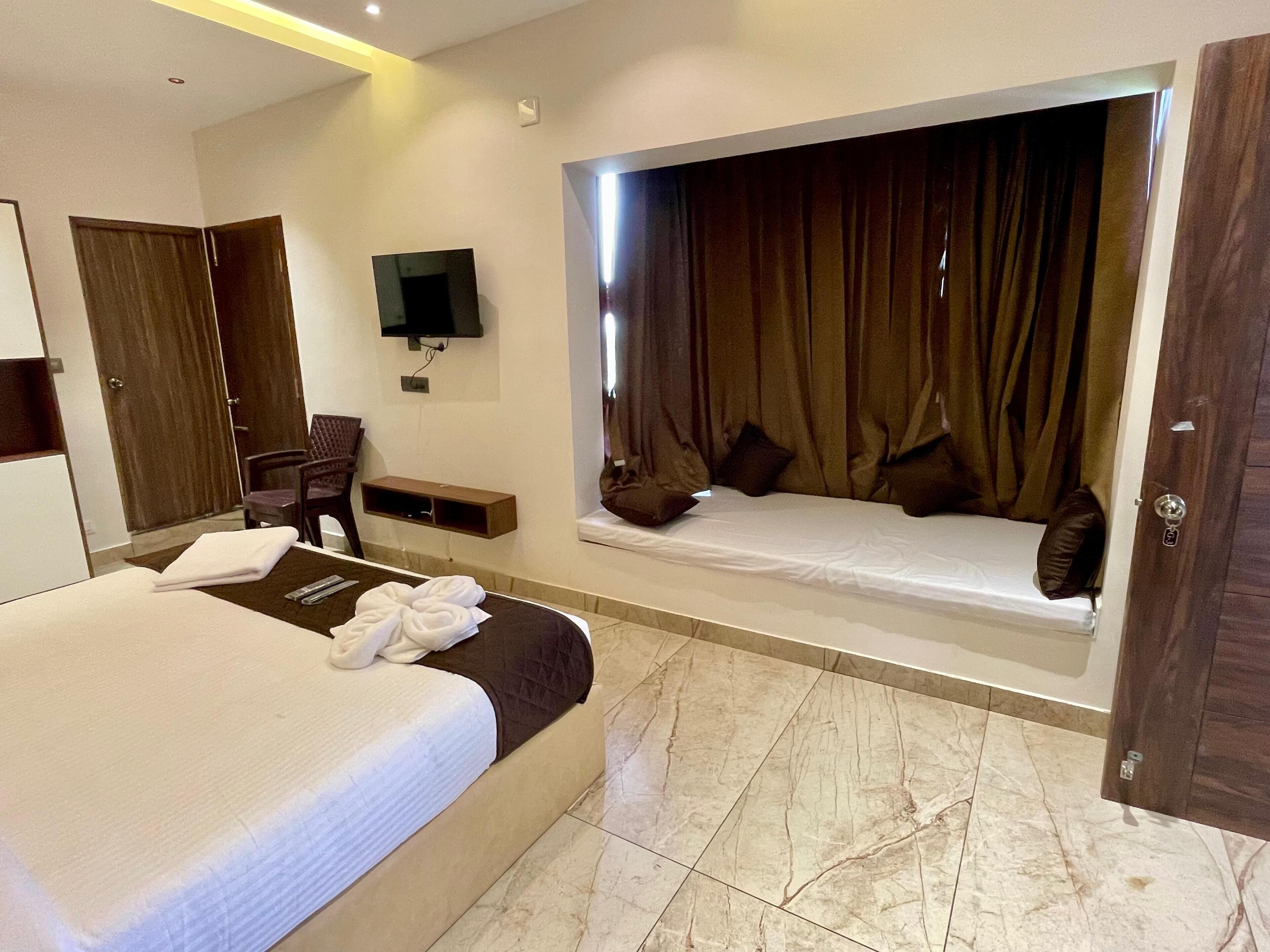 A modern hotel room with a large bed, a seating area with curtains, a flat-screen TV, and a wooden door.