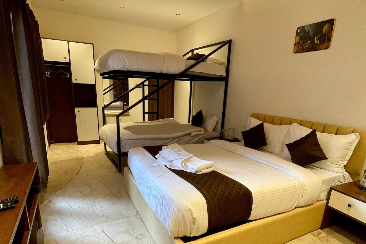 A hotel room with a bunk bed, a single bed, a wooden desk, and a wardrobe.