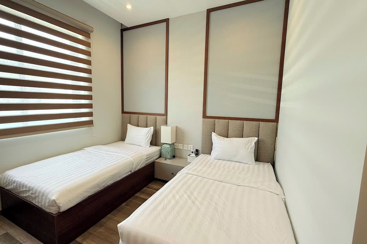 A hotel room with two single beds, a window with blinds, a bedside table with a lamp, and a mirror on the wall.