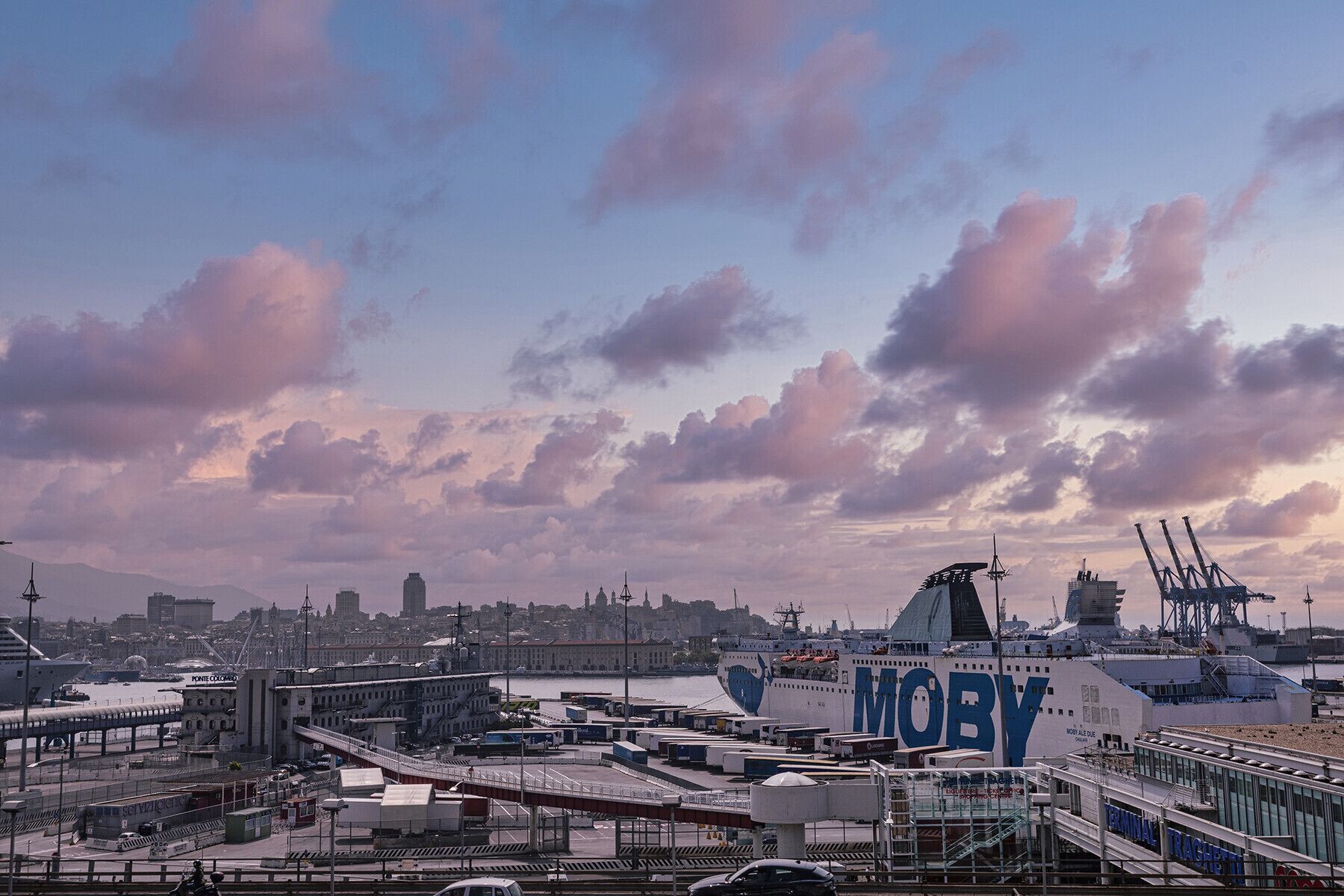 Um porto com um navio grande chamado 'MOBY' atracado, rodeado por edifícios industriais e uma paisagem urbana ao fundo.
