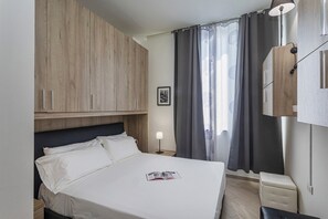 A bedroom with a bed, wooden wardrobe, bedside table, lamp, and a window with curtains. - Portside Genova by Wonderful Italy (Genoa)