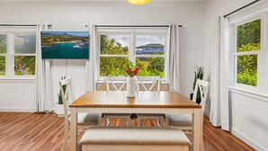 A dining area with a wooden table and chairs, a vase with flowers, and a television displaying a scenic view. - koolkondo 2597 Halalu Guest house (Koloa)