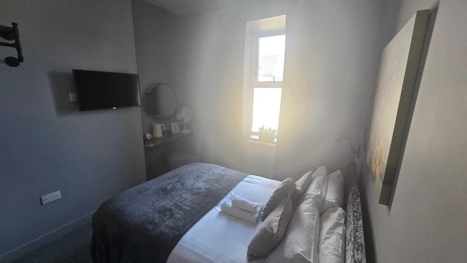 A small, dimly lit room with a single bed, a TV mounted on the wall, and a window providing natural light.