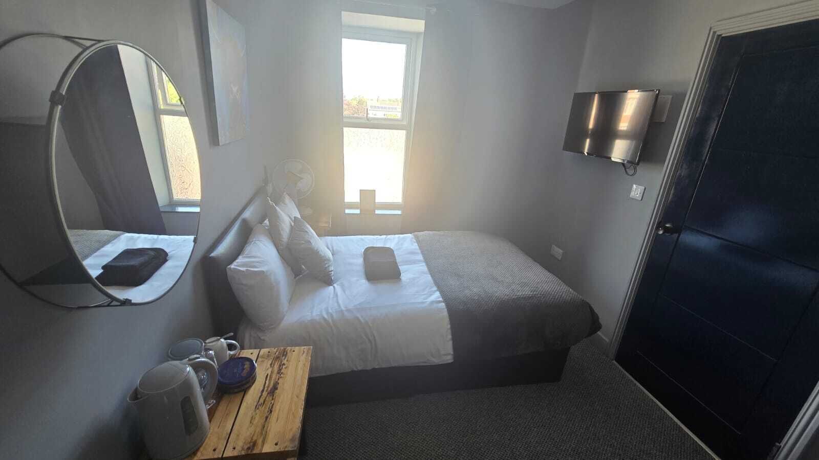 A compact hotel room with a bed, a small table with a cup and a plate, a mirror, a TV, and a door.