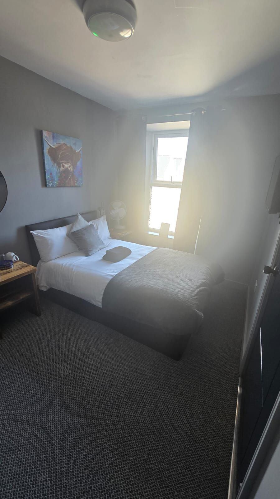 A hotel room with a bed, a nightstand, a painting on the wall, and a window.