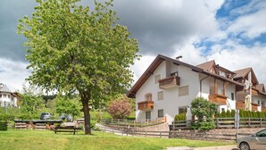 Ein zweistöckiges Chalet mit Balkonen, einer Steinschmalseite und einem grossen Baum im Vordergrund.