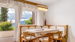 Ein Essbereich mit Tisch, Stühlen, einer Bank, einem Fenster mit Vorhängen, einem Kruzifix an der Wand und Blick ins Freie.