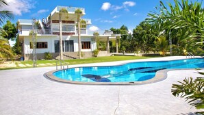 Une maison moderne à deux étages avec une piscine, entourée d’une végétation tropicale.