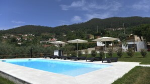 Ein Schwimmbecken mit Liegestühlen und Sonnenschirmen, umgeben von einer Rasenfläche und einer hügeligen Landschaft im Hintergrund.