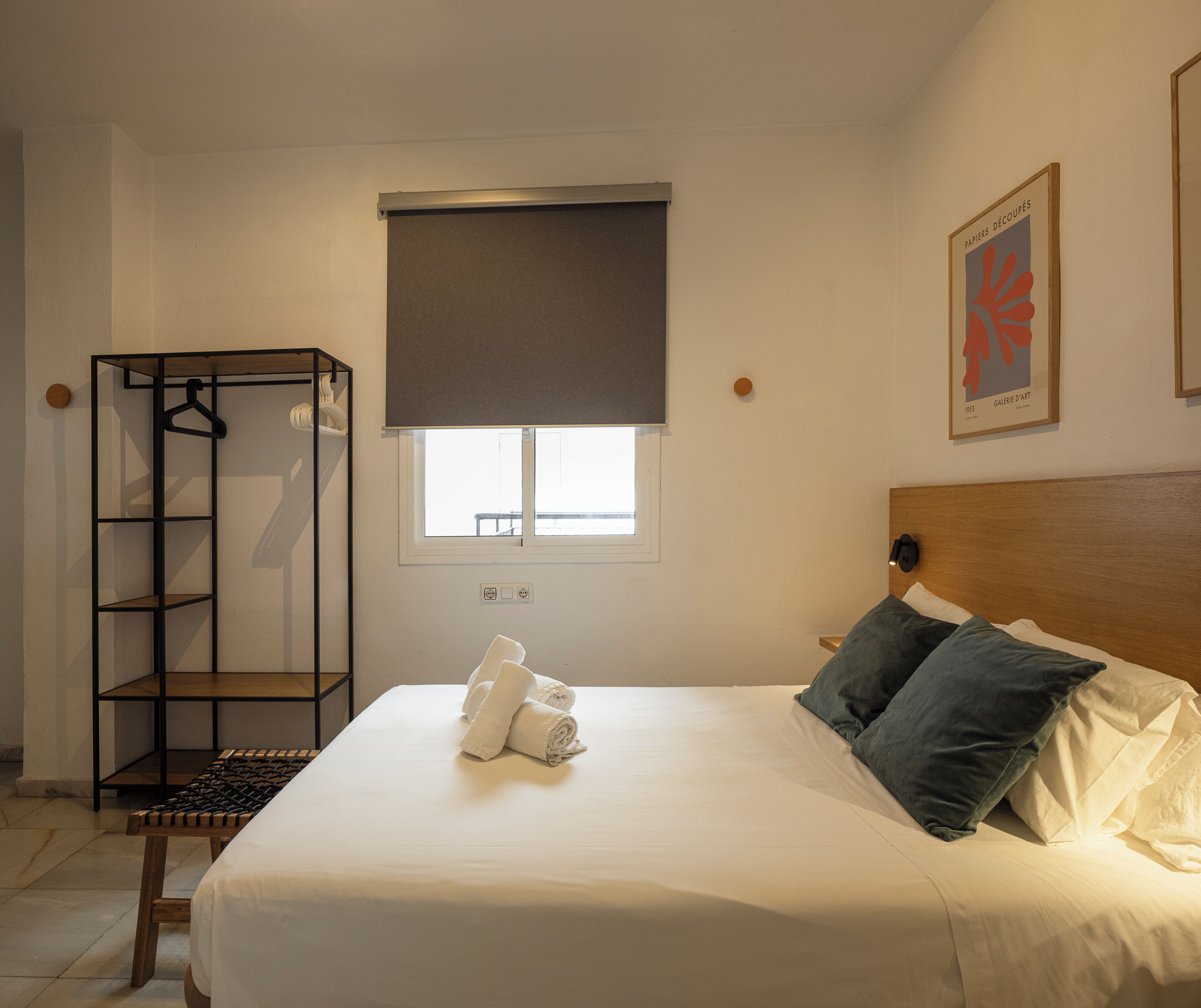 Un dormitorio con cama, mesita de noche, armario de pie, ventana con persiana e imagen en el pared.