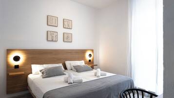 A modern hotel room with a wooden headboard, a large bed with grey bedding, a grey throw blanket, and a small table with a chair.