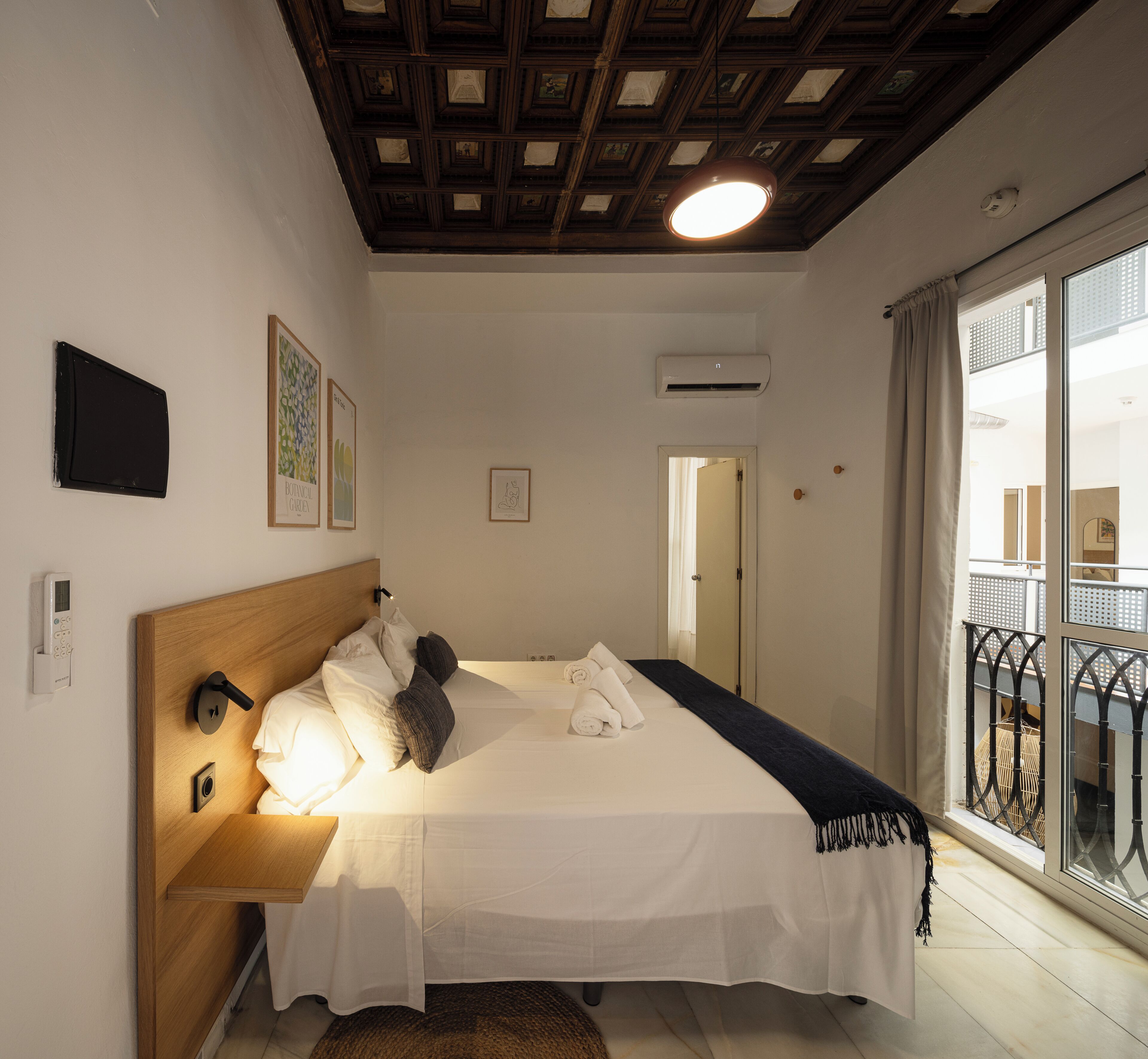 A bedroom with a wooden headboard, a bed with white linens, a bedside table, a wall-mounted air conditioning unit, and a balcony with a metal railing.