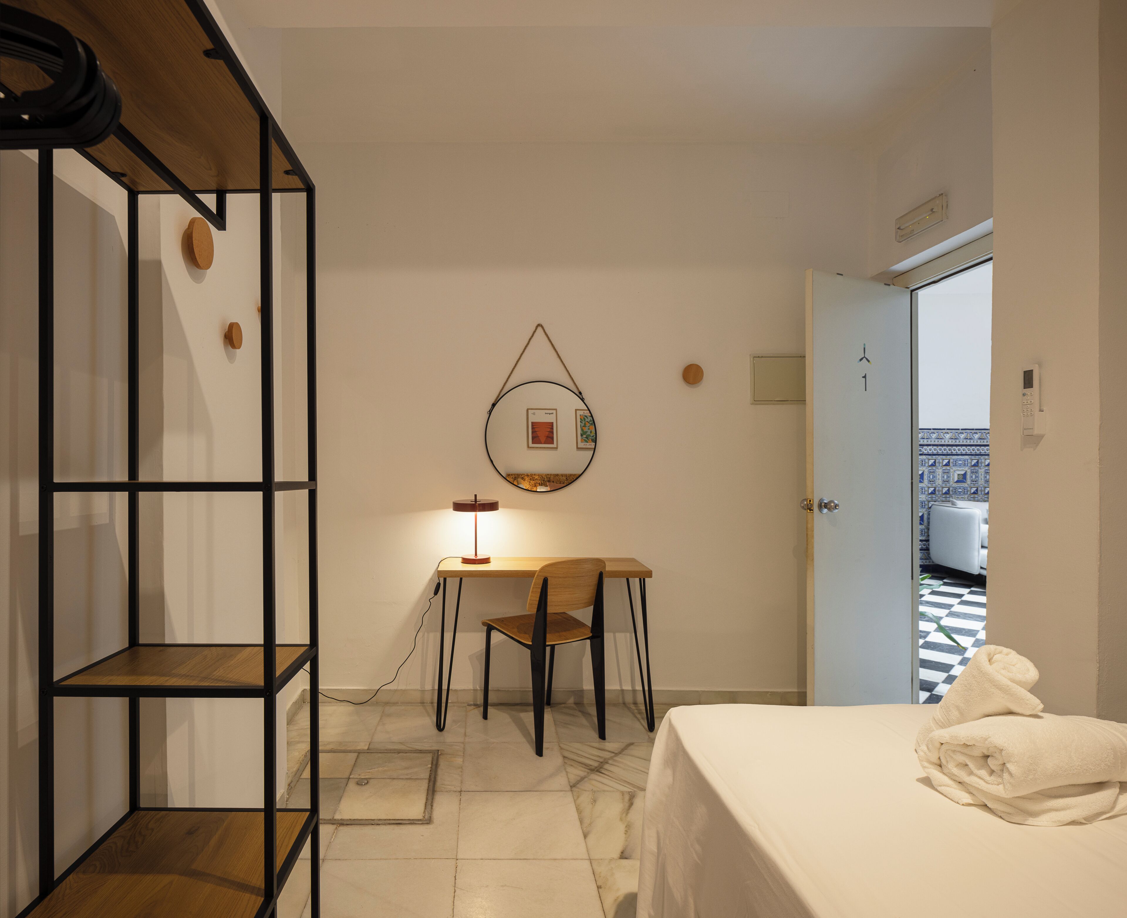 Une chambre d’hôtel comprenant un lit, un bureau avec une lampe, une chaise, une étagère et une porte donnant sur une salle de bain.