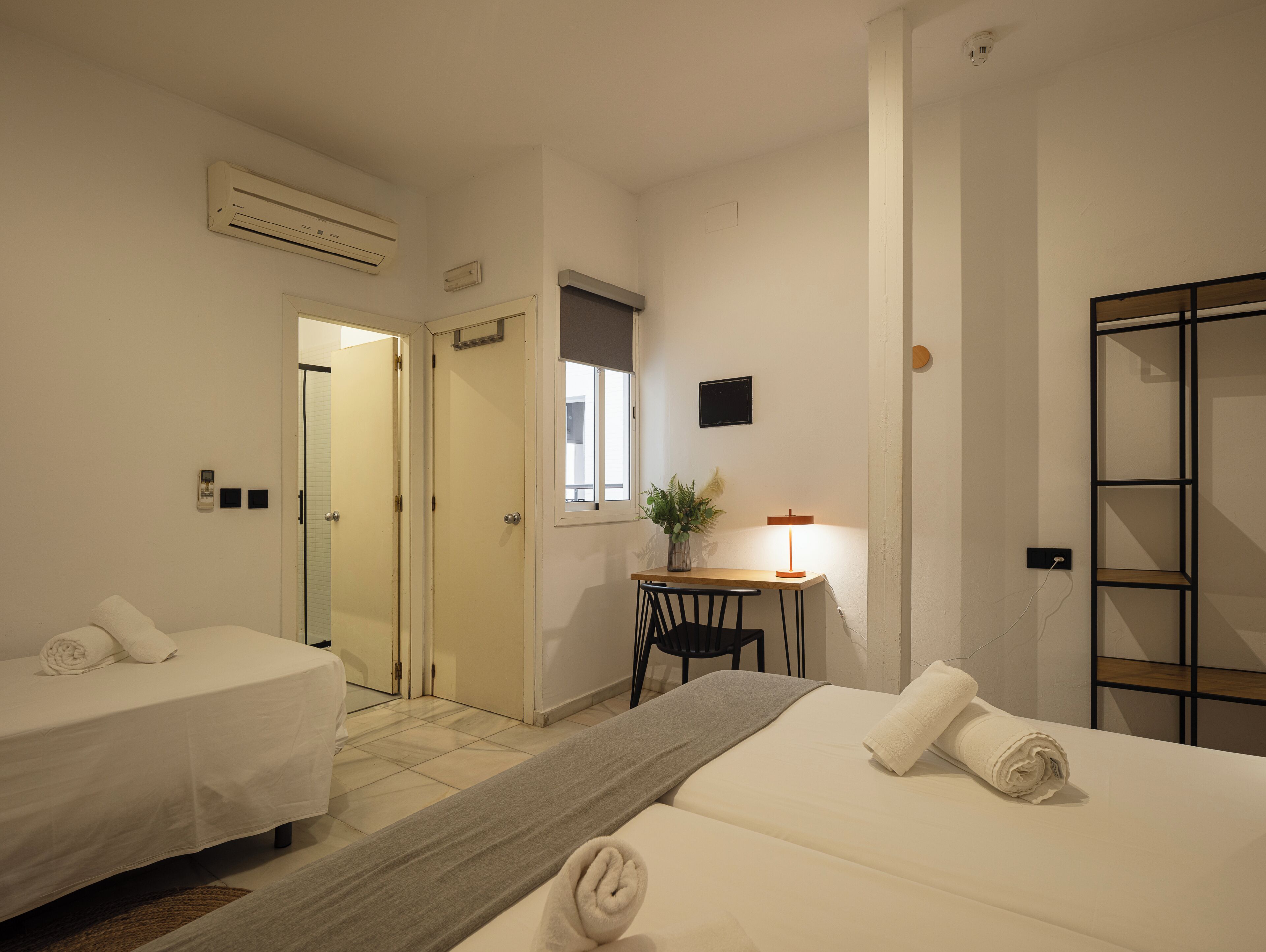 A hotel room with two beds, a desk with a chair, a small table with a lamp, and a window.