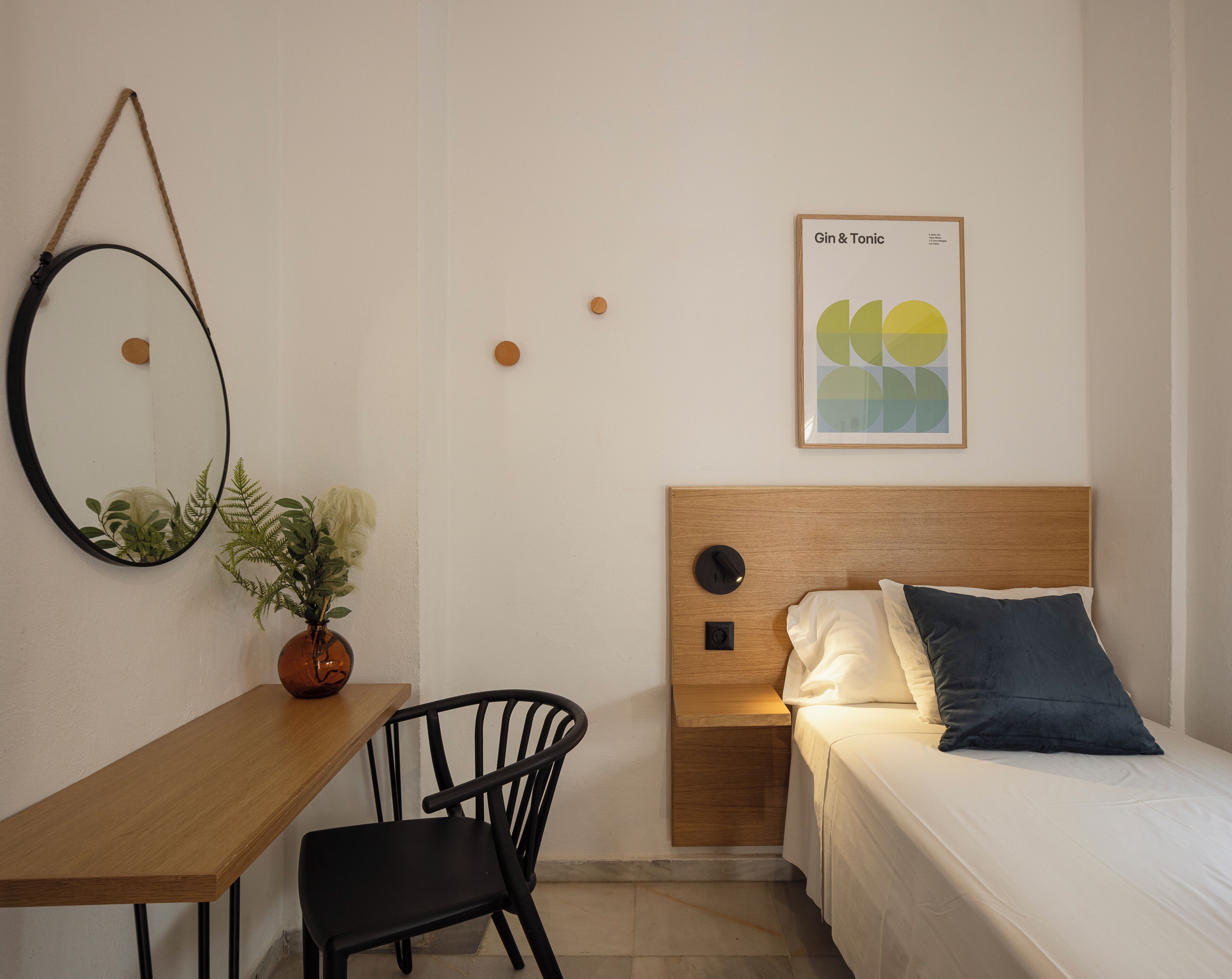A bedroom with a wooden headboard, a bed with white and dark blue pillows, a desk with a chair, a round mirror, and a framed picture on the wall.