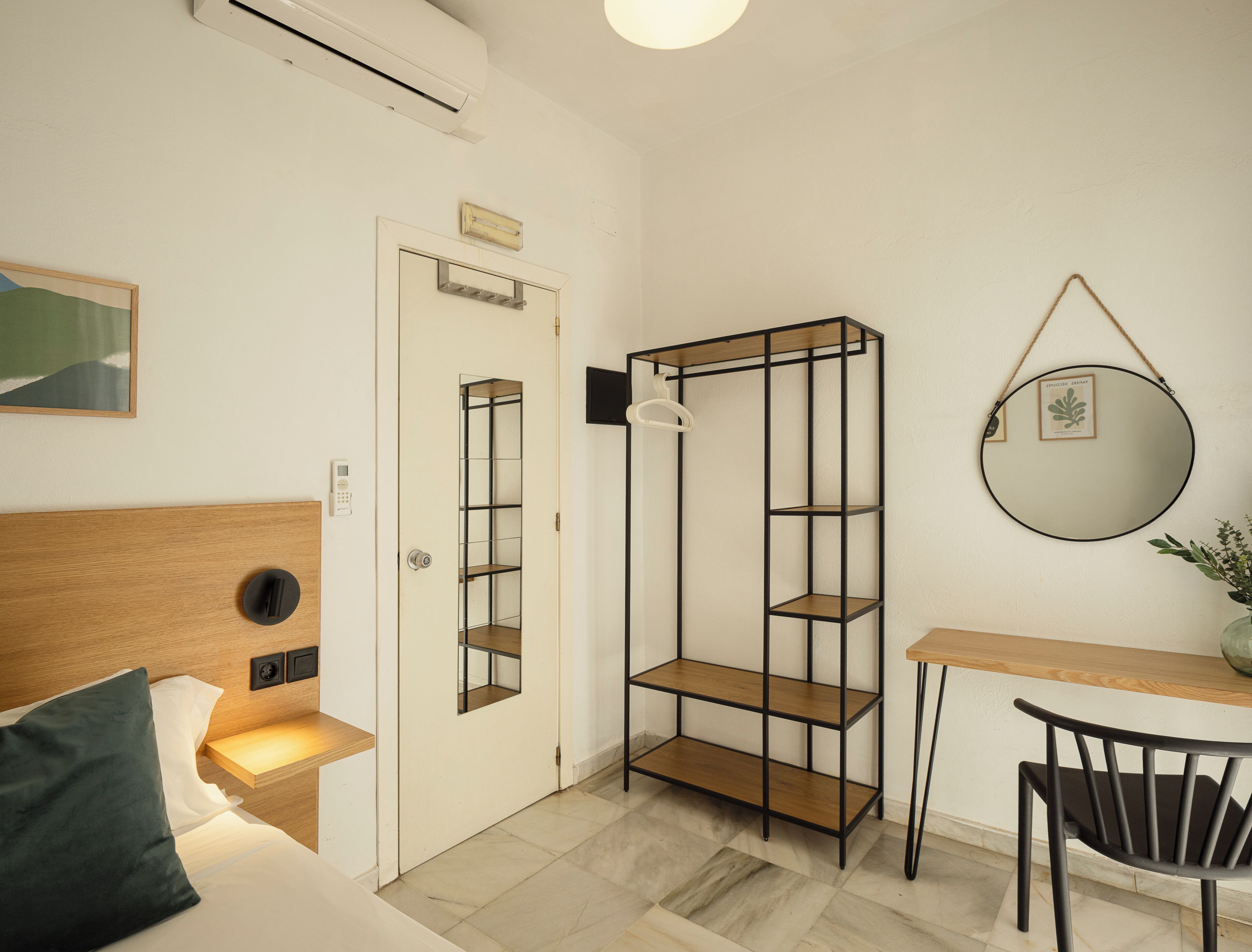 A compact room with a bed, bedside table, a wall-mounted mirror, a small shelf unit, and a desk with a chair.