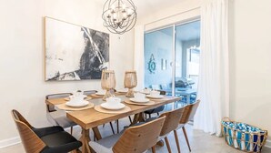 A dining area with a wooden table set for four, surrounded by chairs with wooden frames and black cushions. A chandelier hangs above, and a large abstract painting is on the wall.
