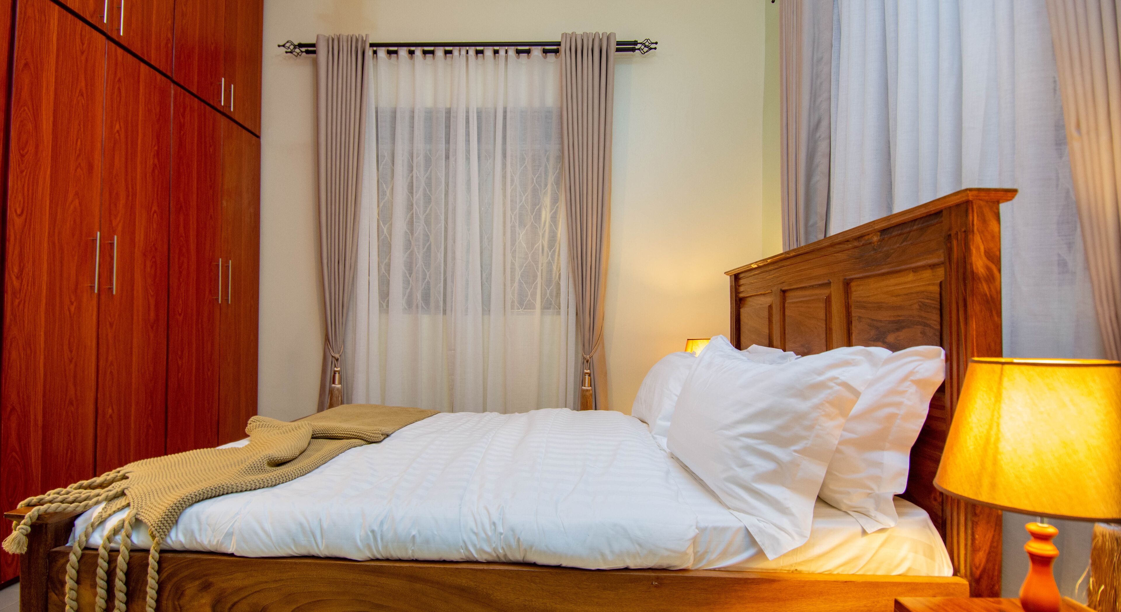 A neatly made bed with white linens and a beige throw blanket, a wooden headboard, and a bedside lamp.