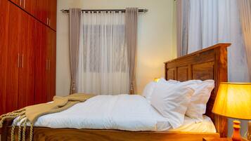 Een netjes opgemaakt bed met witte lakens en een beige plaid, een houten hoofdbord en een bedlampje.