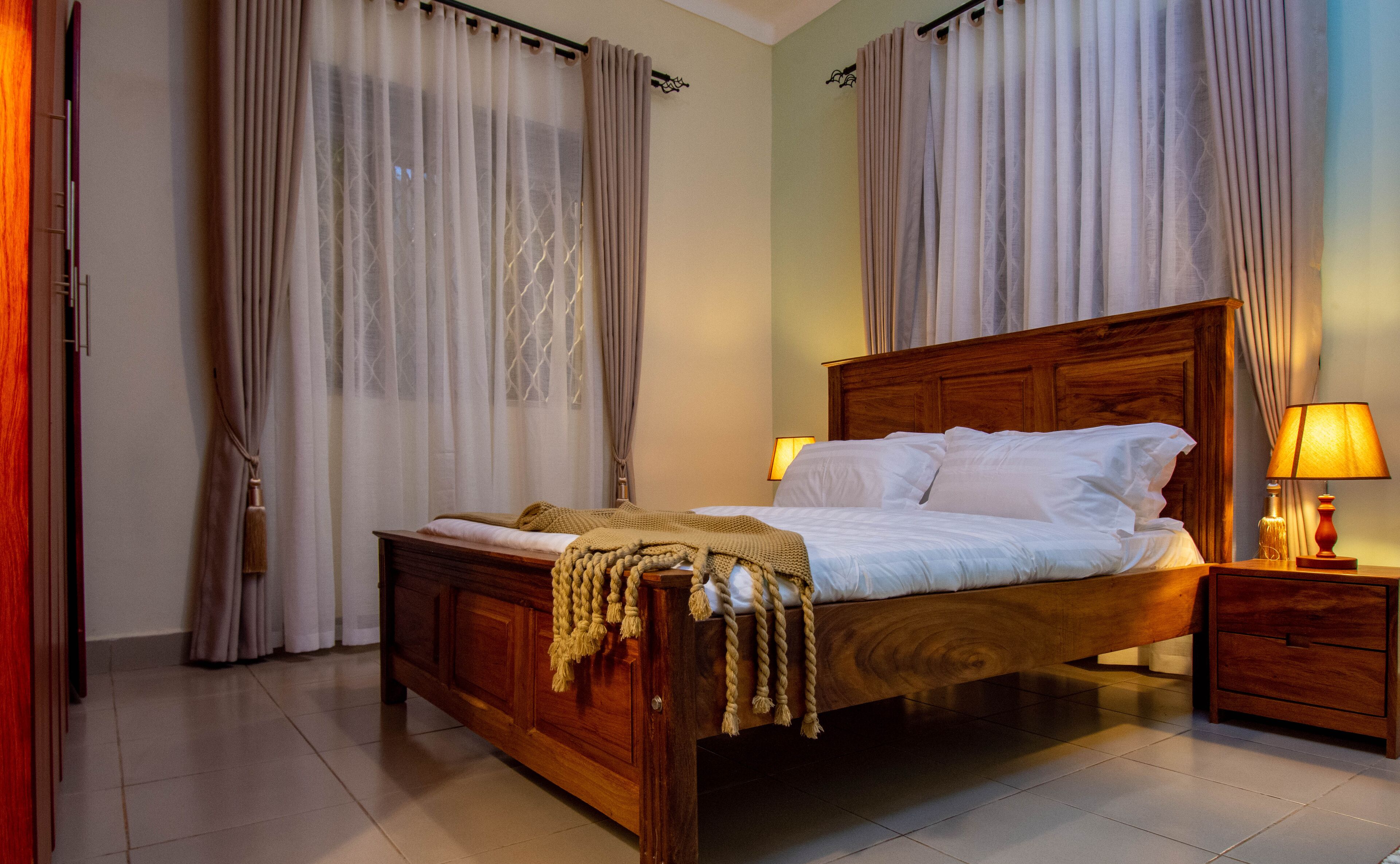 A neatly made bed with a wooden headboard, a bedside table with a lamp, and a window with sheer curtains.