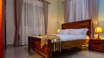 A neatly made bed with a wooden headboard, a bedside table with a lamp, and a window with sheer curtains.