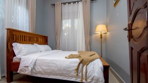 A neatly made bed with white linens and a beige throw blanket, a wooden headboard, and a floor lamp with a beige shade. - Urban Oasis (Kampala)