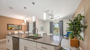 A modern kitchen with a central island, a dining area with a blue chair, and a ceiling fan.