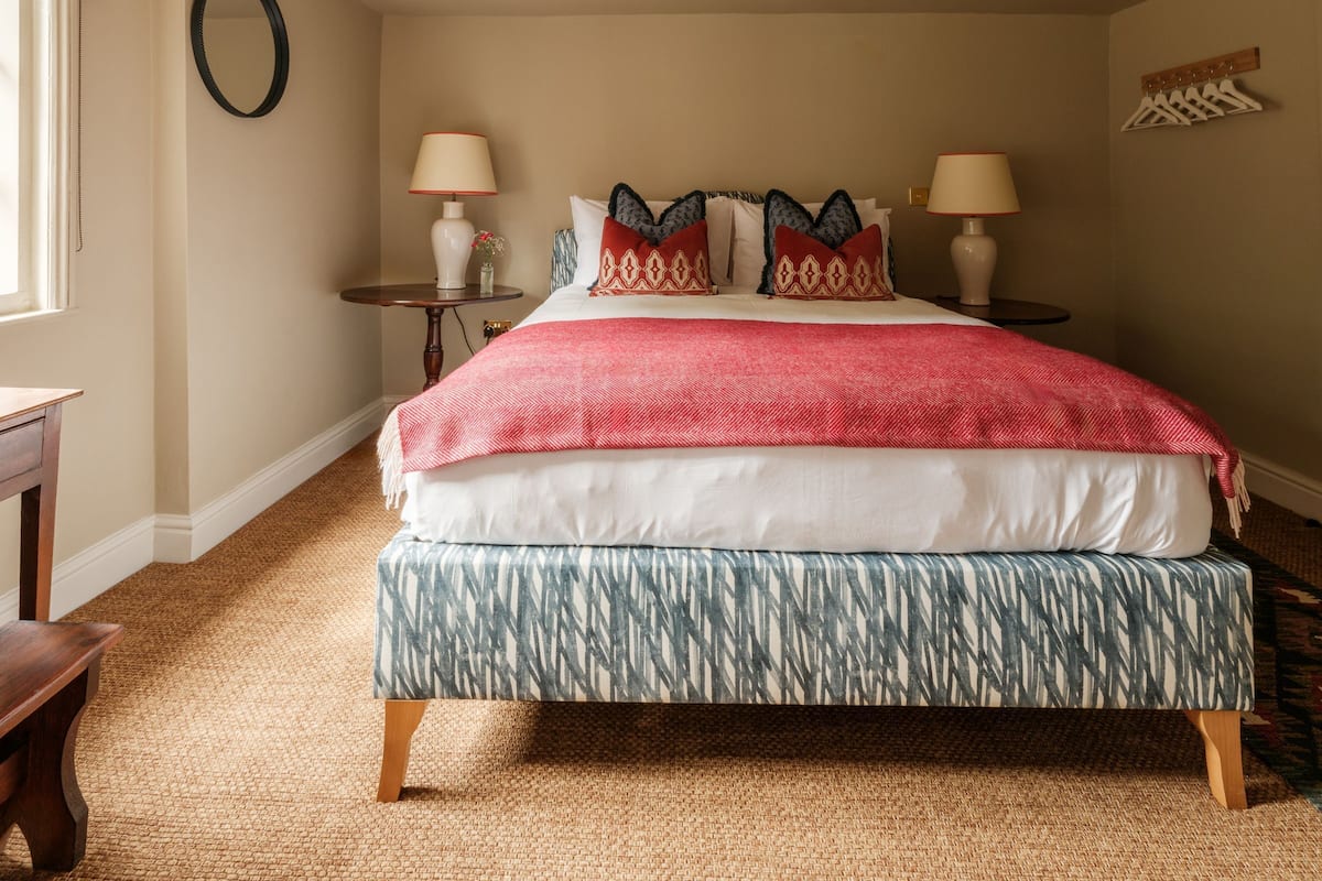 A neatly arranged bedroom with a large bed, two bedside tables with lamps, a round mirror on the wall, and a small table with a vase.