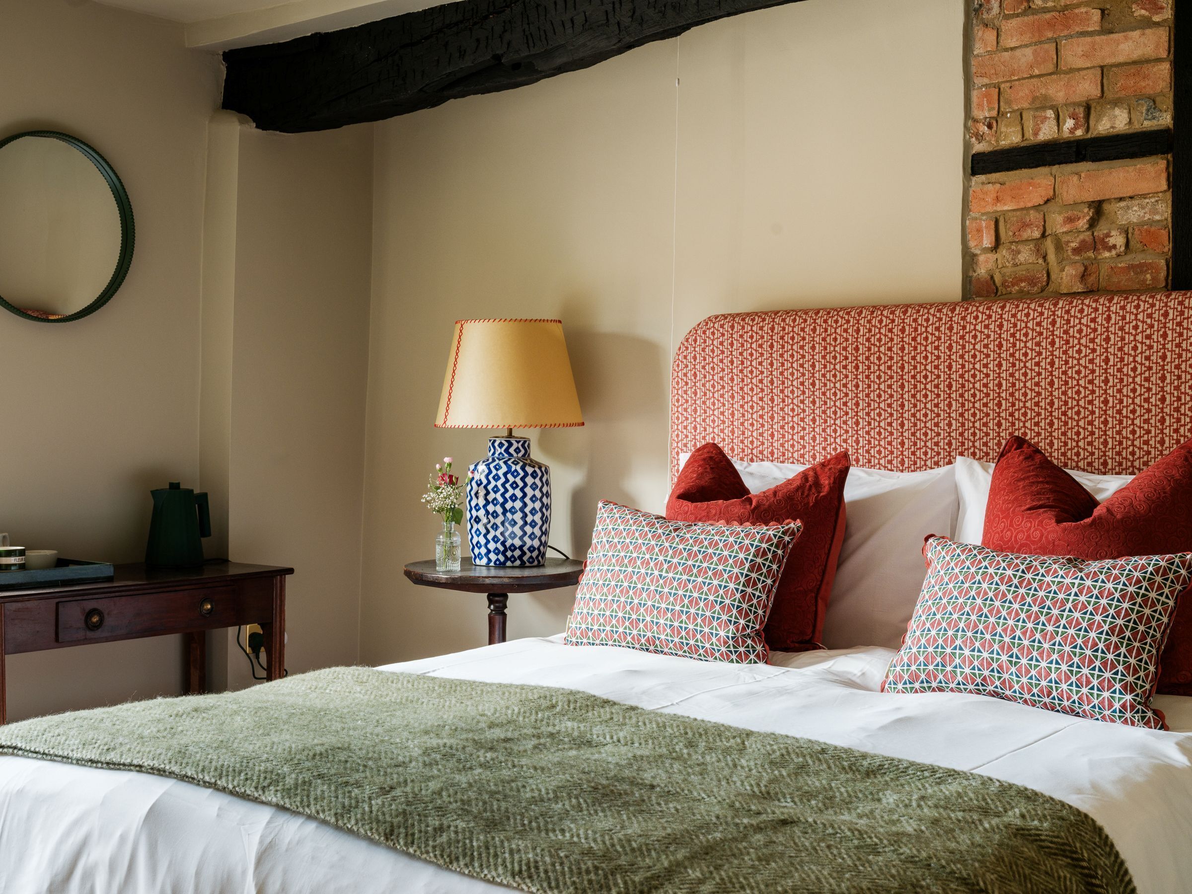 A neatly made bed with a green blanket, a patterned vase with flowers, and a lamp on a bedside table.