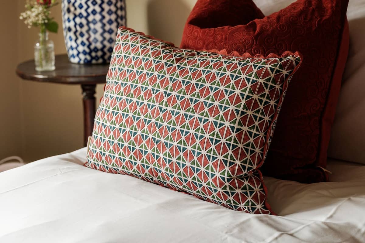 A neatly made bed with patterned pillows, a lamp, and a vase with flowers on a bedside table.