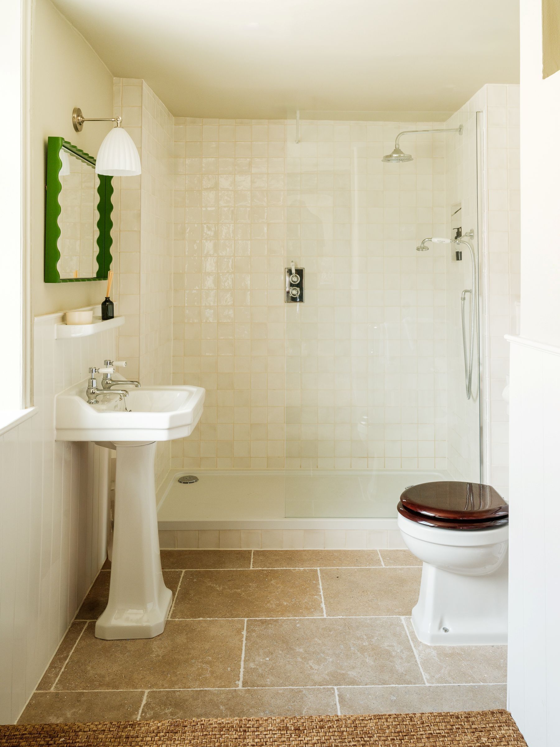 A bathroom with a pedestal sink, a toilet with a wooden seat, a shower enclosure, and a mirror with a green frame.