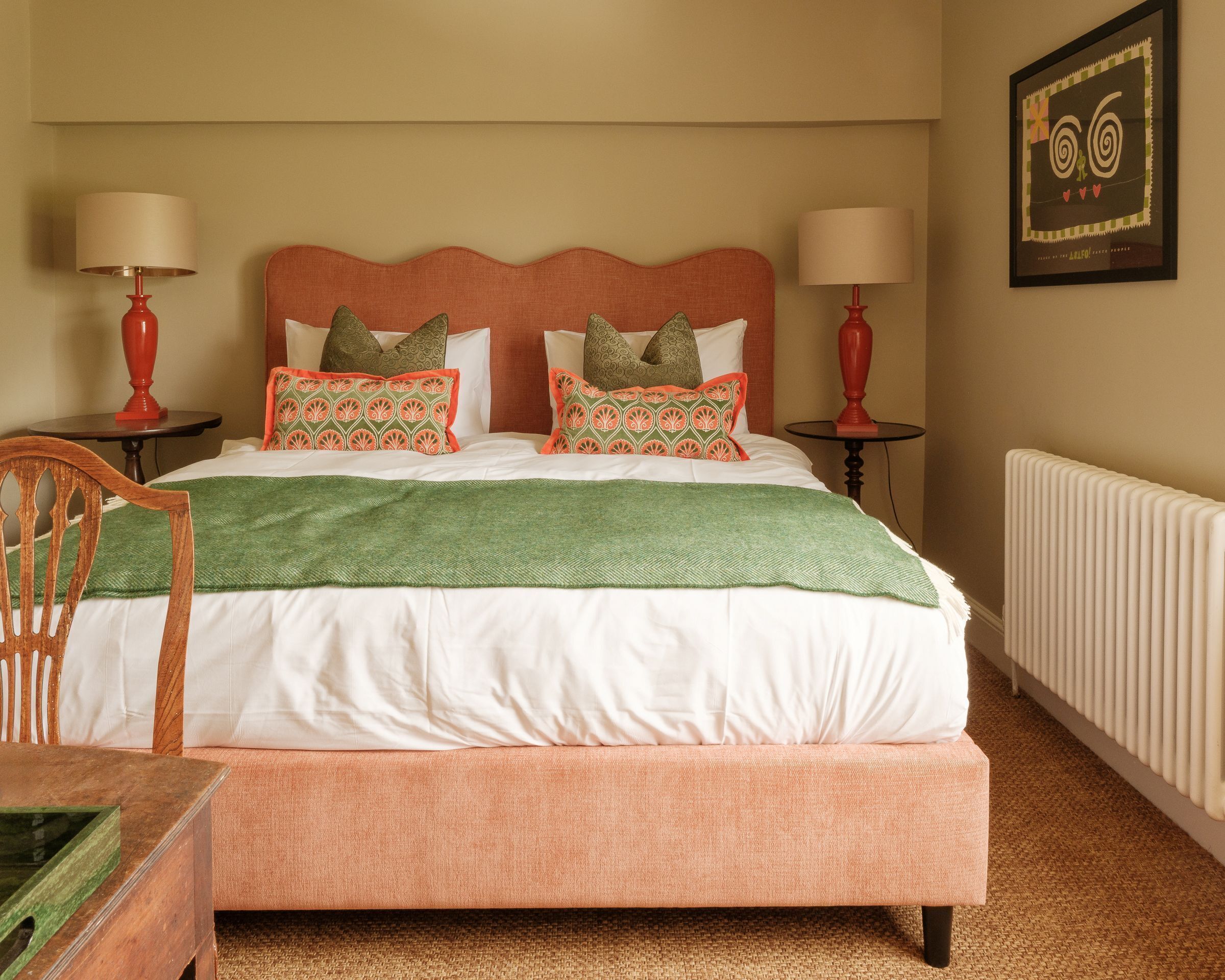 Una cama bien hecha con un plaid verde, dos mesitas de noche con lámparas, un cuadro enmarcado en la pared y un radiador.