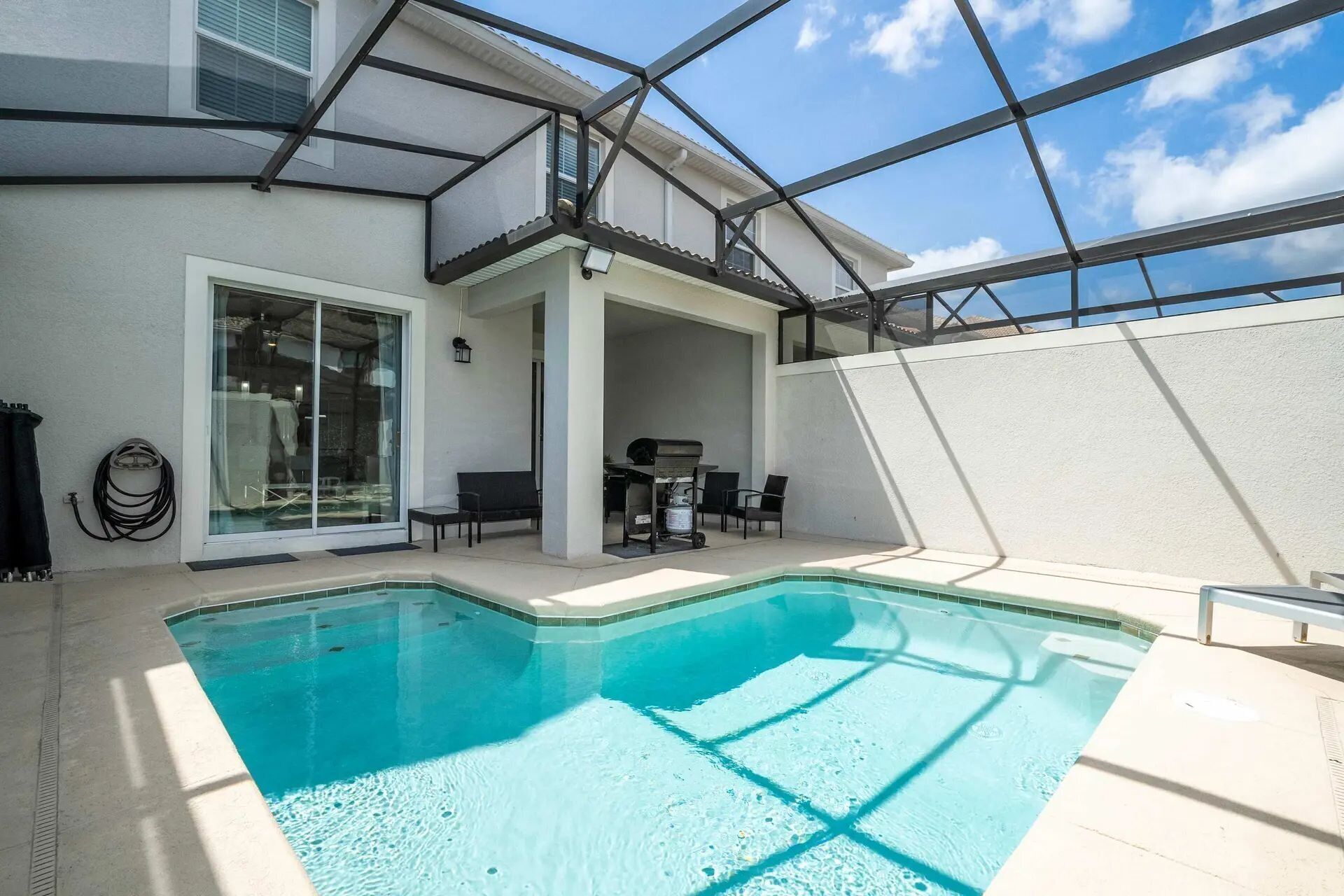 A modern outdoor pool area with a glass-enclosed patio, a barbecue setup, and a clear blue sky.