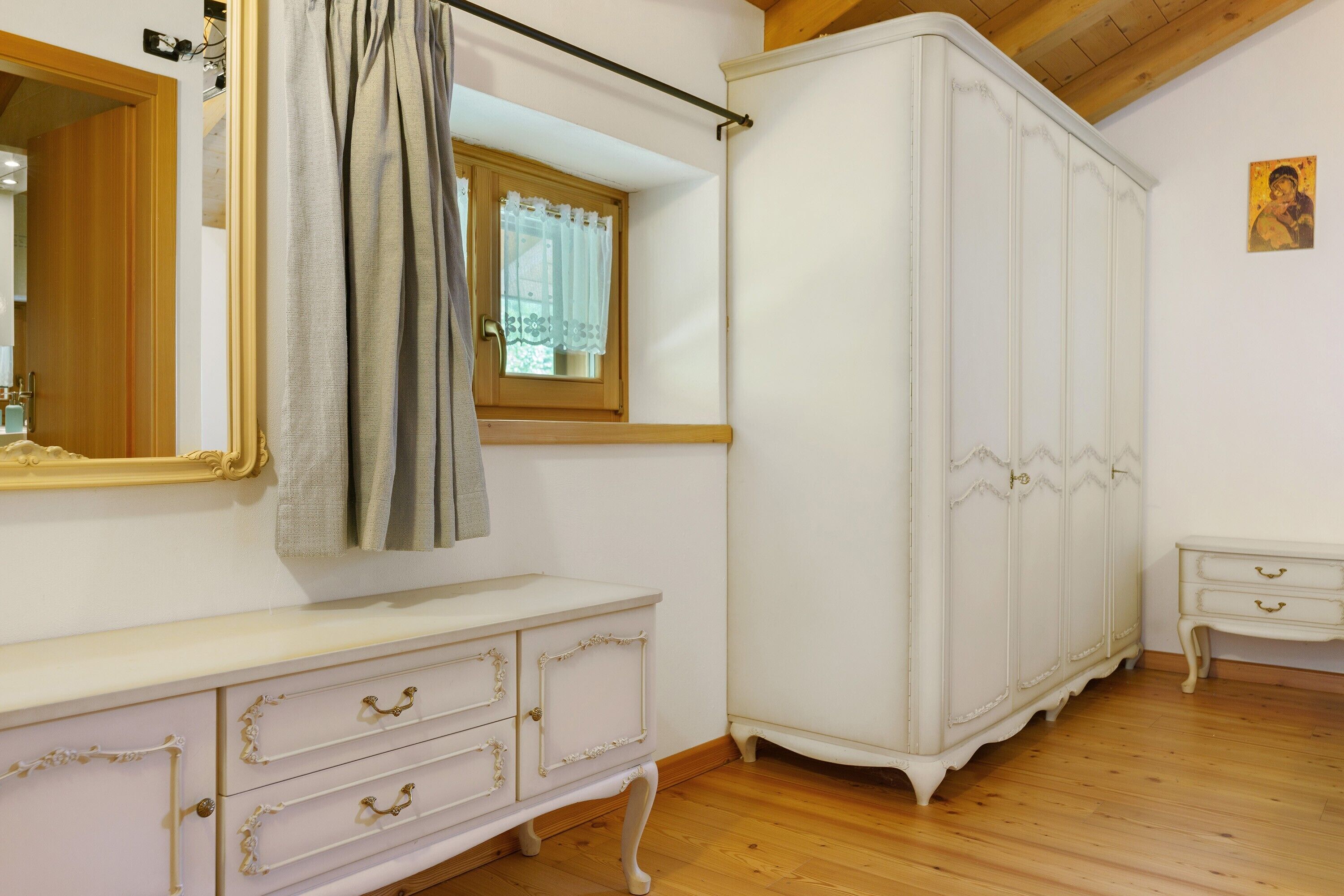 A room with a wooden floor, a white wardrobe, a white dresser with drawers, a mirror, and a window with a curtain.