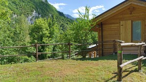 Eine Holzhütte mit einer Holzterrasse und einem Metallgeländer, umgeben von üppiger Vegetation und Bergen.