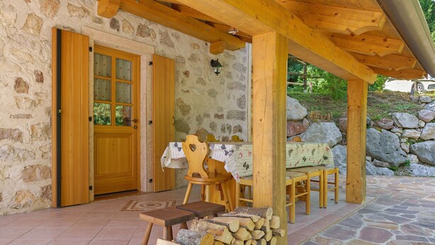 Ein Steinhaus mit einer Holztür, einer überdachten Terrasse mit Tisch und Stühlen und einem Holzstapel.