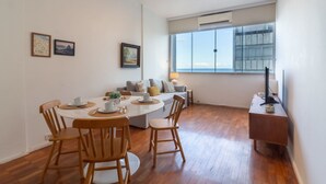 Un comedor con una mesa redonda, sillas de madera y un sofá. Hay cuadros en la pared, una ventana con persianas y una mesita con una lámpara.