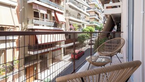 A balcony with wicker chairs and a glass table, overlooking a street with parked cars and multi-story buildings. - Spacious Luxury Apartment | Near Piraeus Port (Piraeus)