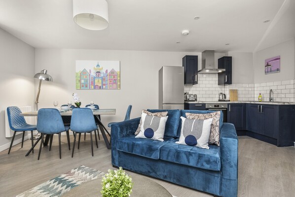 A modern living room with a blue sofa, dining table, and chairs. There is a kitchen area with dark cabinets and a white tiled backsplash. - Buzzing Brighton Apartments | By My Getaways (Brighton)