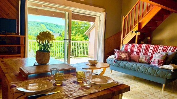 Una habitación acogedora con una mesa de madera lista para comer, un sofá azul con cojines estampados y vistas a un paisaje verde exuberante a través de una puerta corrediza de vidrio.