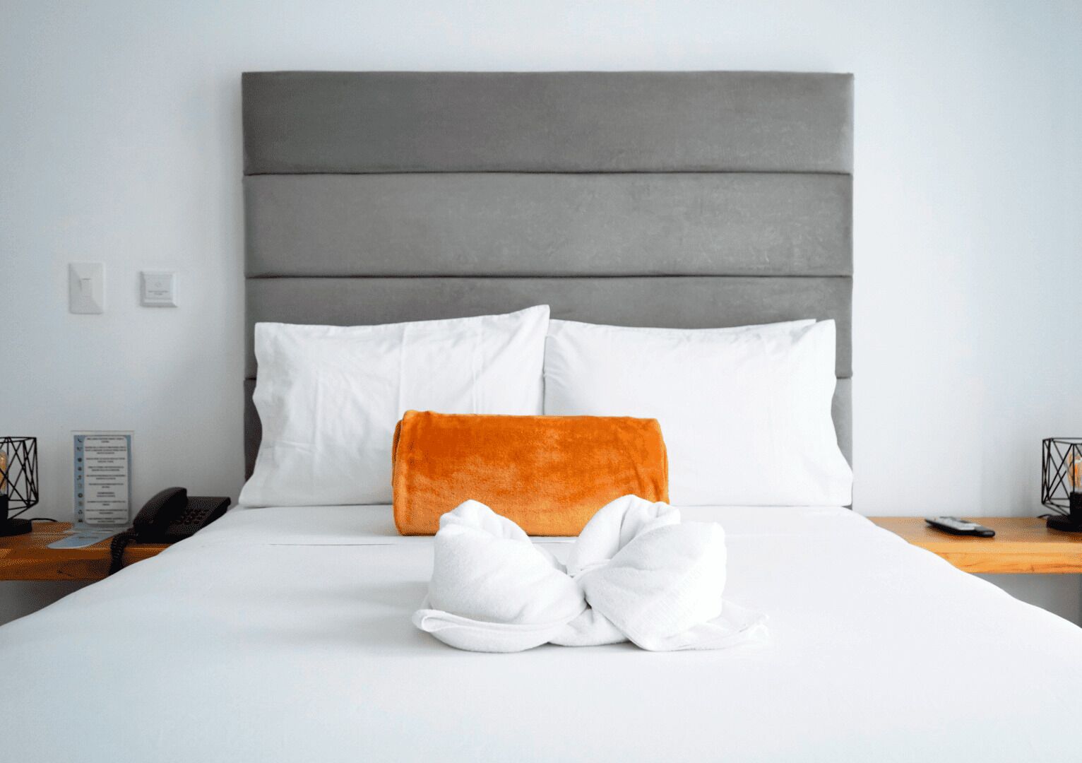 A neatly made bed with white and orange pillows, a grey headboard, and a bedside table with a phone and a candle holder.